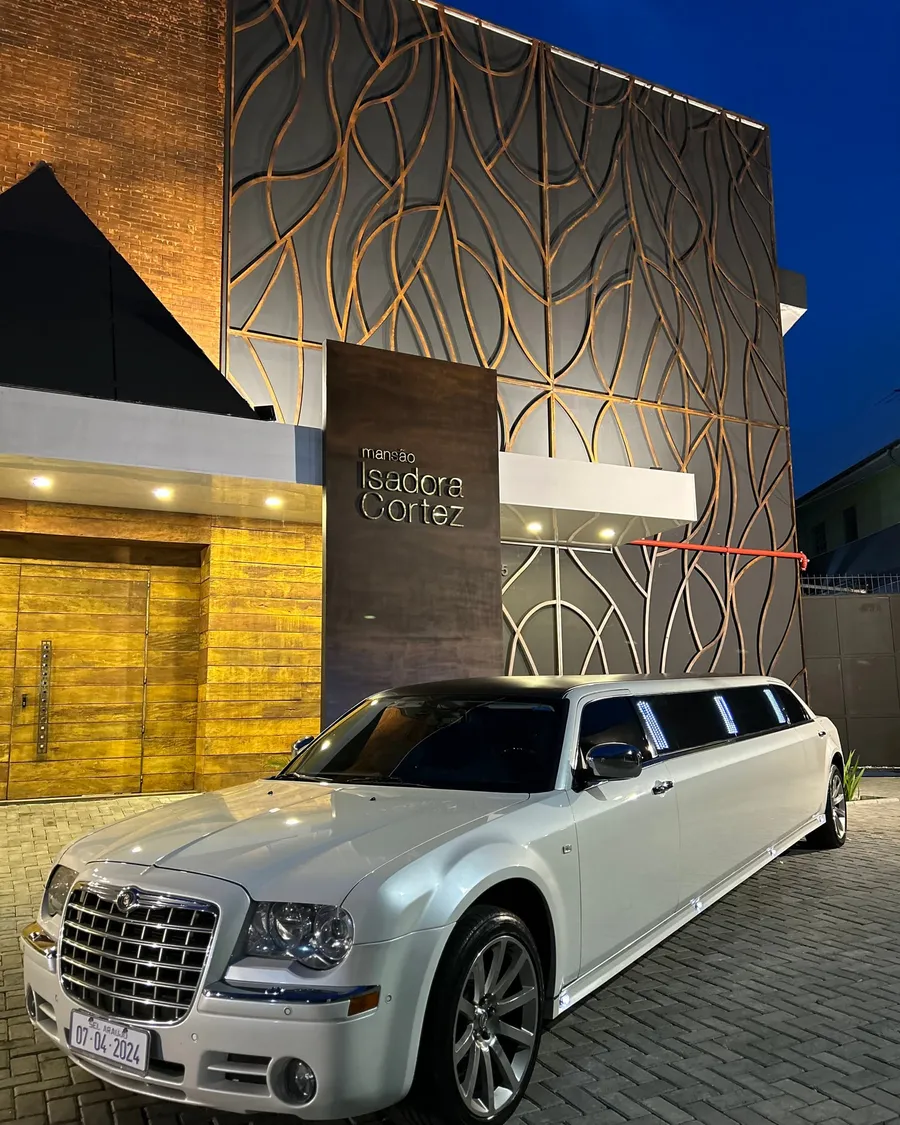 Limousine Chrysler 300C branca em três quartos, parada na entrada de um salão de eventos ao anoitecer