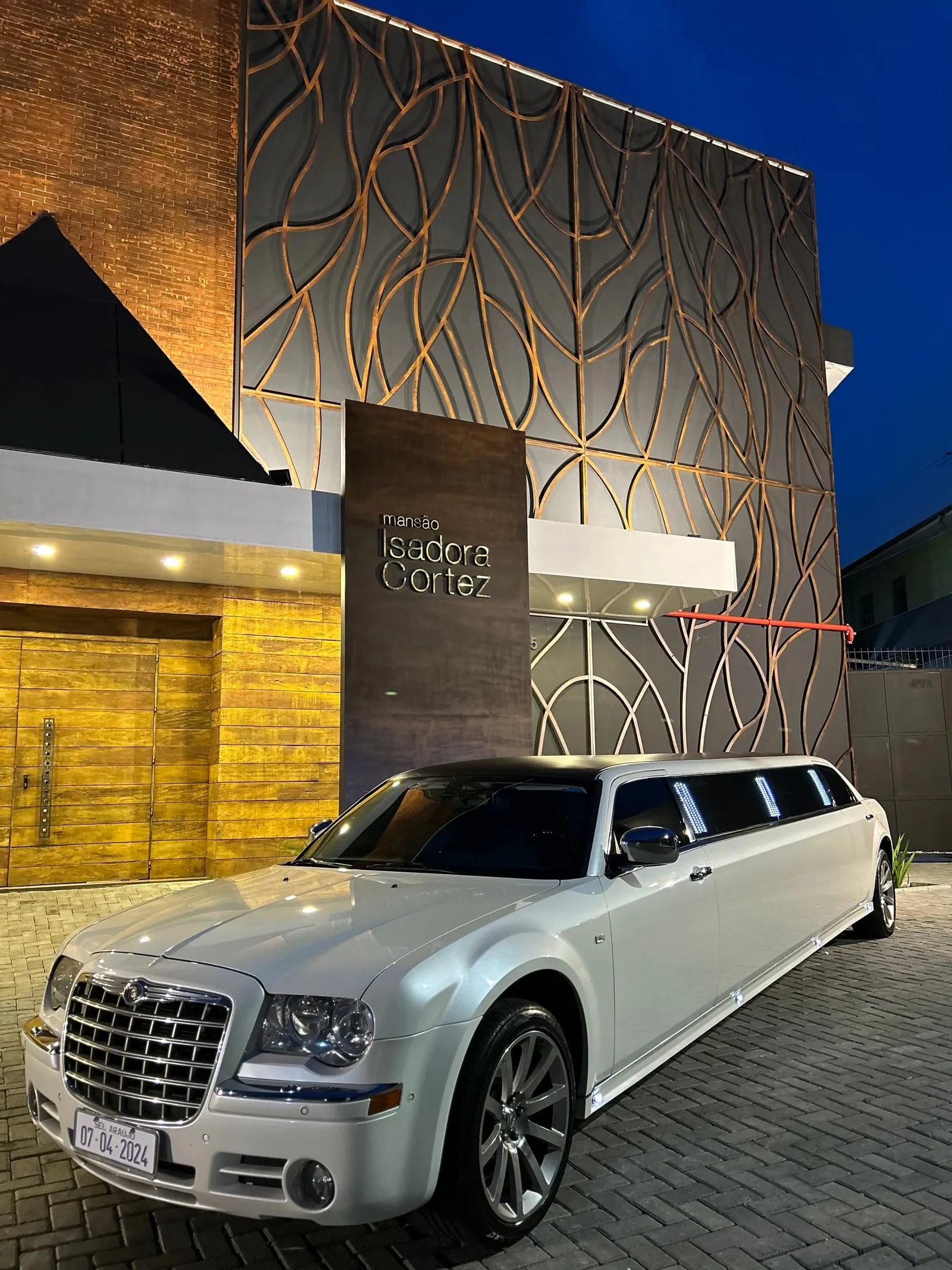 Limousine Chrysler 300C branca em três quartos, parada na entrada de um salão de eventos ao anoitecer